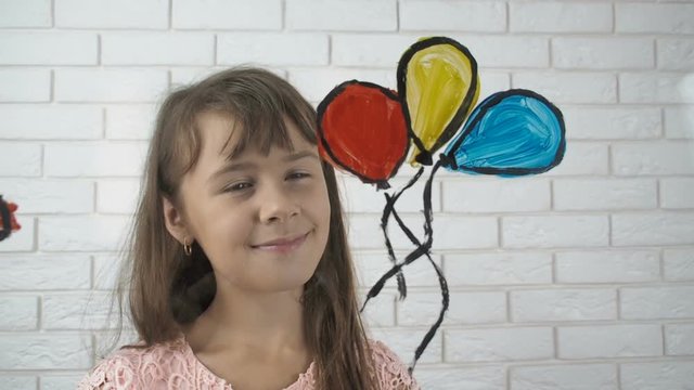 The child draws balloons and a flying kite. A little girl paints inflatable balls.
