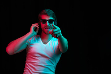 Enjoying his favorite music. Happy young stylish man in sunglasses with headphones listening sound and smiling while standing against blue neon background