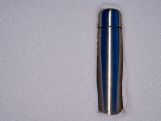 A large silver metal thermos with tea stands in a snowdrift. winter evening forest. copy space
