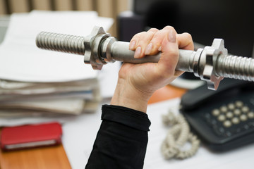 Female hand holding a dumbbell on the desktop background. In the background-working documents, phone and computer. Concept: sports during the break.
