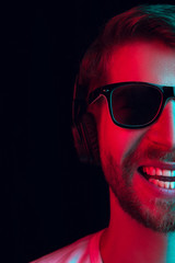 Fototapeta premium Enjoying his favorite music. Happy young stylish man in sunglasses with headphones listening sound and smiling while standing against blue neon background