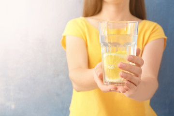 Young woman holding glass of lemon water on color background, closeup