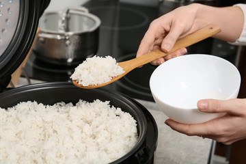 Woman putting boiled rice into bowl from cooker in kitchen, closeup