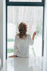 Back view of curly girl standing in front of window with coffee cup