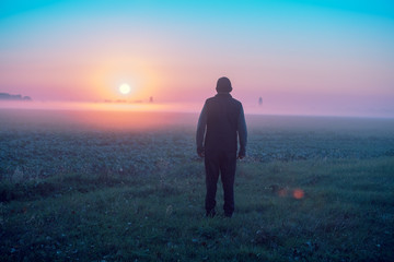 A man stands in the field in the early morning and looks at the sunrise