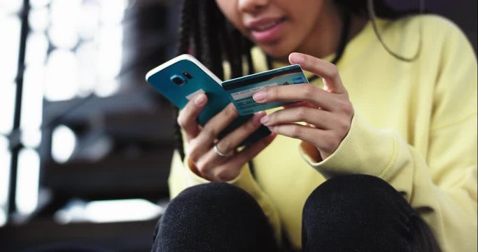 Details closeup african lady order something from her smartphone using a bank card she very careful typing her bank details , she sitting down on the modem stairs in the office. 4k