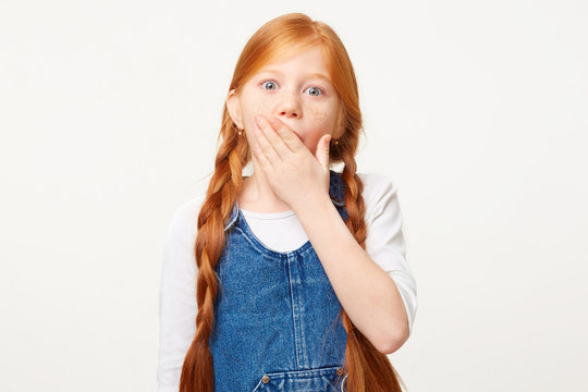 Portrait of scared kid in panic nervous dressed in denim overall keeps palm on mouth, feals fear, isolated on white background. Red-haired girl commited offense. OMG concept