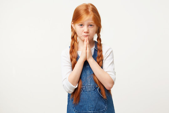 Red-haired kid with pleading facial expression hoping wishing something, folded arms in a prayer gesture. Girl with braids want a special gift, stands against white background.