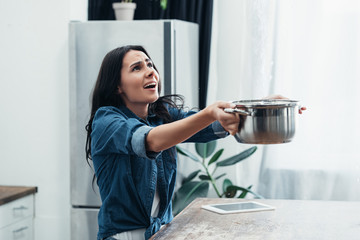 Worried woman in denim short with pot dealing with water damage in kitchen