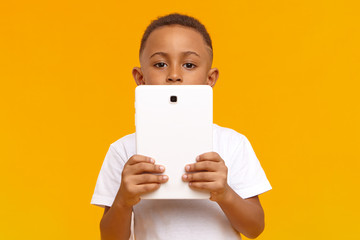 Picture of smart dark skinned schoolboy holding generic white digital tablet, doing homework, looking for information on internet. Cute curious African American male child using touch pad pc