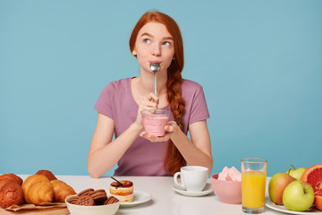 Nice red-haired girl with a braid sits at a table, has lunch, holds in her hands a glass, dreamingly looks at left upper corner and licks teaspoon with pleasure after tasting yummy yogurt.
