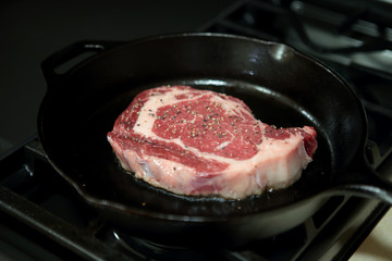 Ribeye steak frying in a cast iron pan on a natural gas stove top.