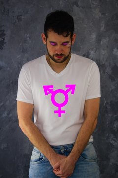 A man with make up in t-shirt with third gender symbol- transgender.The man looks like a woman. He stands and has folded hands in front of his crotch.He is ashamed and sad.