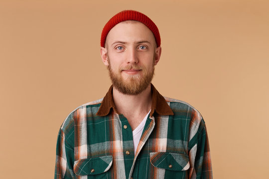 Attractive young man in studio looking at camera. Portrait of a normal guy with broad in red hat and checkered shirt in front of beige background