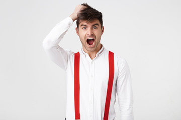 Indoor photo of stupefied astonished man wears shirt and red suspender, keeps hand on head,...