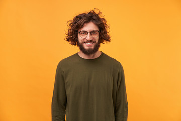 Cute friendly bearded man in glasses with curly hair smiling looks happy isolated on yellow background. A good friend came to visit, a niceguy.