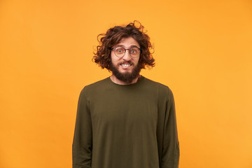 A man with a beard in glasses and curly dark hair looking at the camera, bite lip, confused bewildered as if made mistake, feel guilty isolated on a yellow background dressed in ordinary clothes. - obrazy, fototapety, plakaty