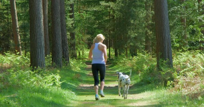 A mature woman running through a sunny forest with her pet dog.