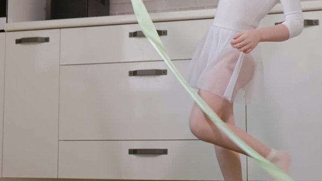 A happy little girl in a white gymnastic swimsuit trains, dances with a ribbon for rhythmic gymnastics, jumps and performing professional exercises.