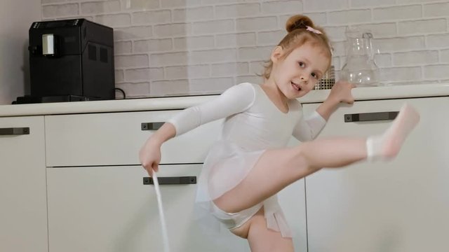 A happy little girl in a white gymnastic swimsuit trains, dances with a ribbon for rhythmic gymnastics, jumps and performing professional exercises.