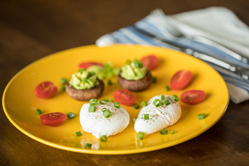 Poached eggs with avocado stuffed mushrooms, tomatoes, and green onions on a cheery yellow plate