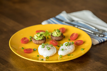 Poached eggs with avocado stuffed mushrooms, tomatoes, and green onions on a cheery yellow plate