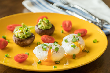 Poached eggs with avocado stuffed mushrooms, tomatoes, and green onions on a cheery yellow plate