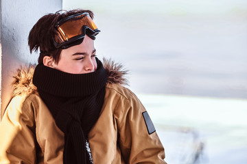 Portrait of a teenage snowboarder dressed in snowsuit and protective goggles sitting on railing near the snowy beach