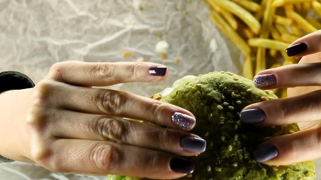 Women's hands with a beautiful manicure hold a big juicy burger with a meat patty and a green bun. Women's hands spin a burger in their hands and bring it to their mouths against the background of the