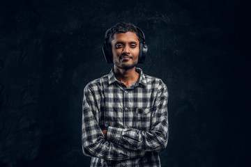 Handsome Indian guy is happy listening to music in wireless headphones standing with his arms crossed in studio against the background of the dark wall