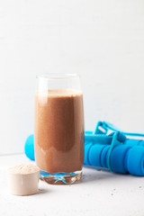 Glass of Chocolate Protein Shake with milk and banana, Whey protein in scoop and dumbbells. Sport nutrition. Bright stone / wooden background. Copy space. 