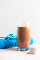 Glass of Chocolate Protein Shake with milk and banana, Whey protein in scoop and dumbbells. Sport nutrition. Bright stone / wooden background. Copy space. 