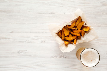 Fried potato wedges in paper box, glass of beer on a white wooden surface, top view. Space for text.