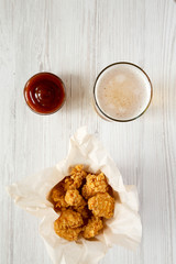 Fried chicken bites in paper box, barbecue sauce, glass of beer over white wooden surface, top view. Flat lay, overhead, from above.