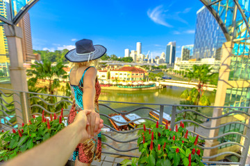 Fototapeta premium Follow me. Tourist woman with wide hat holding hand of partner looking panorama from terrace over Clarke Quay and Riverside area in Singapore, Southeast Asia. Lady enjoys on Singapore River downtown.