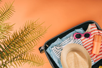 top view of suitcase with summer accessories and sunscreen on orange background with palm leaves