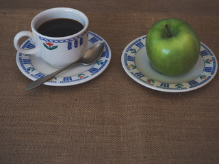 Breakfast of a porcelain coffee cup, an old spoon and a plate with a green apple on a vintage...