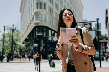 female asian tourist holding paper map searching direction standing on road on sunny day in city urban san francisco. young college girl finding tourist spot famous attraction in travel trip usa.