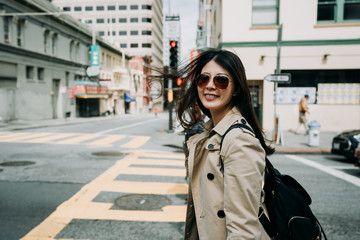 beautiful cheerful lady backpacker walking on zebra cross turning head face camera smiling. young girl with sunglasses standing on road in busy street san francisco usa in summer. elegant asian woman