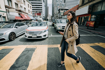 full length of young asian office lady walking on zebra cross going to work. girl backpacker holding cell phone searching online map in busy city urban. cars vehicle waiting stop traffic light