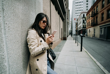 young elegant office lady relying on wall standing in quiet street in busy city urban in san francisco usa. asian woman using mobile phone chatting online waiting near building in cloudy day.