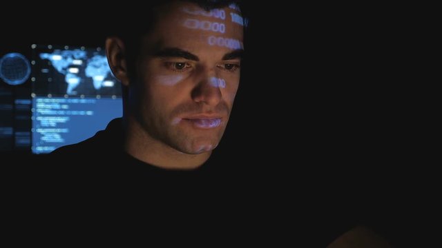 Man programmer hacker working on a computer while blue code characters reflect on his face in a dark office room.