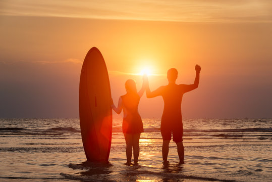 Silhouette Of Surfer Man With Young Girl Standing Happily On The Beach Looking Out To The Sea And Raise Their Hand In The Sunset After Surfing.