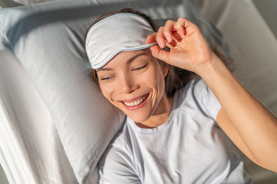 Happy Asian Woman Waking Up Feeling Refreshed From Beauty Sleep Eye Sleeping Mask For A Good Night's Rest. Smiing Young Biracial Girl In Bed. Healthy Sleep Pillow Comfort.