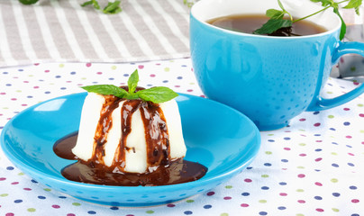 Cream pudding with chocolate sauce, mint cup of tea on multicolored tablecloth.