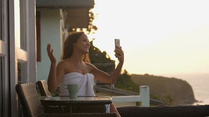 Young woman having video chat showing view sharing travel experience from hotel balcony at sunset using smart phone connecting with mum on social media summer vacation during sunset