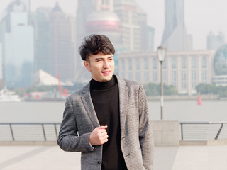 Portrait of a handsome Chinese young man in casual suit smiling and looking away confidently with Shanghai bund background, winter fashion, cool young man lifestyle. 