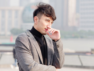 Naklejka premium Portrait of a handsome Chinese young man in casual suit thinking with hand on chin with Shanghai bund background, winter fashion, cool young man lifestyle.