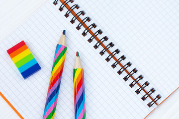 Open copybook with two rainbow-coloured pencils and a rubber on white wooden table