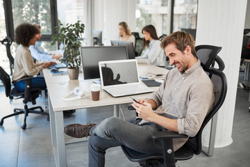 CEO with smile sitting in his chair and using smart phone. Office interior, in background employees working.